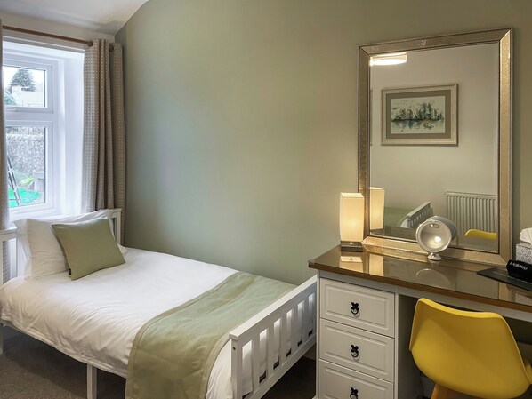 Un dormitorio con una cama, una mesita de noche con lámpara, un espejo, una silla y una ventana con cortinas.