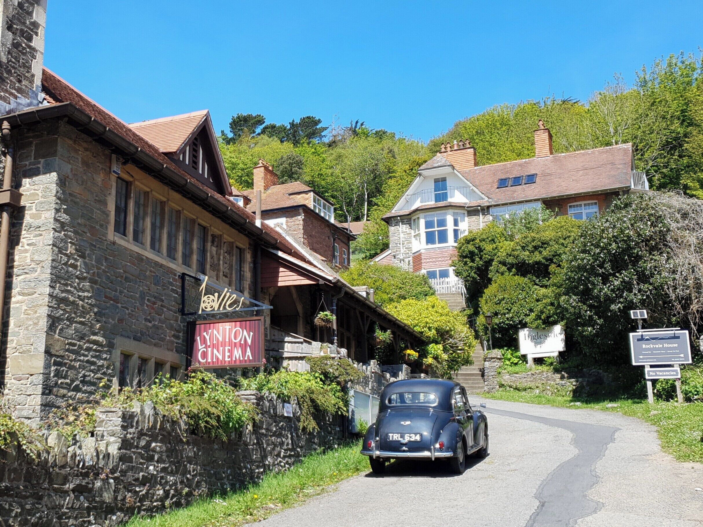Une voiture ancienne est garée devant un cinéma à la façade en pierre, sur laquelle on peut lire l’inscription « Lynton Cinema ».