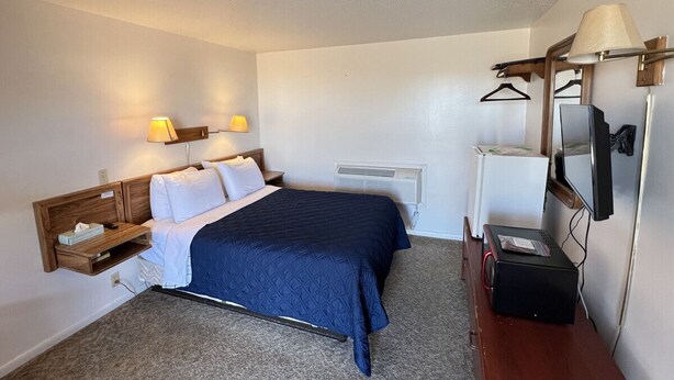 A hotel room with a bed, bedside lamps, a TV, a heater, and a desk with a computer. - Room 102 - Standard Queen - Sleeps 2 - West Lake Grandvue Inn (Downs)