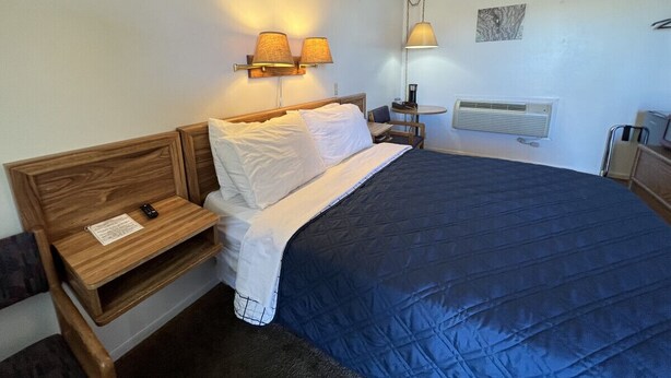 A neatly made bed with a blue quilt, a wooden nightstand with a lamp, and a wall-mounted heater.