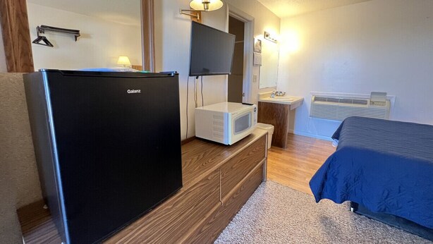 Ein Hotelzimmer mit schwarzem Kühlschrank, einer Mikrowelle, einem Flachbildfernseher, einem Nachttisch mit Lampe, einem Bett mit blauer Bettdecke, einer Holzkommode und einem Teppichboden.