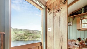 A wooden door with a view of a river and hills, a coat rack with hooks, and a mirror reflecting a room with a fur rug and a tray of food. - Keepers Hide - UK43676 (Oldfield, near Keighley)