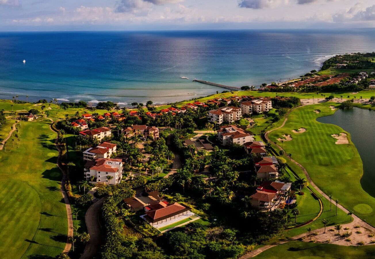 An aerial view of a coastal resort with multiple buildings, a golf course, and a body of water.
