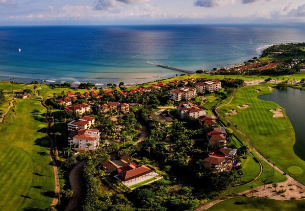 An aerial view of a coastal resort with multiple buildings, a golf course, and a body of water.