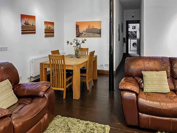 A living room with a brown leather sofa, a wooden dining table with chairs, and a green rug. There are three paintings on the wall.