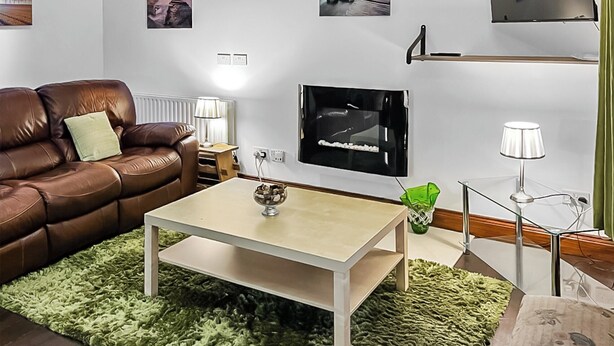 Un salon avec un canapé en cuir marron, une table basse blanche, un tapis à poils verts, un téléviseur à écran plat et une petite plante verte.