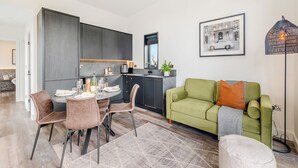 Una cocina moderna con armarios oscuros, una zona de comedor con una mesa redonda y sillas, un sofá verde y una lámpara de pie.