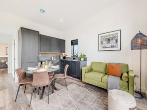 A modern kitchen with dark cabinets, a dining area with a round table and chairs, a green sofa, and a floor lamp.