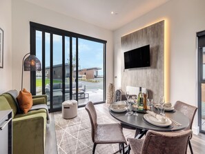 Un comedor moderno con una mesa preparada para cuatro personas, un sofá, una lámpara de pie y un televisor montado en la pared.