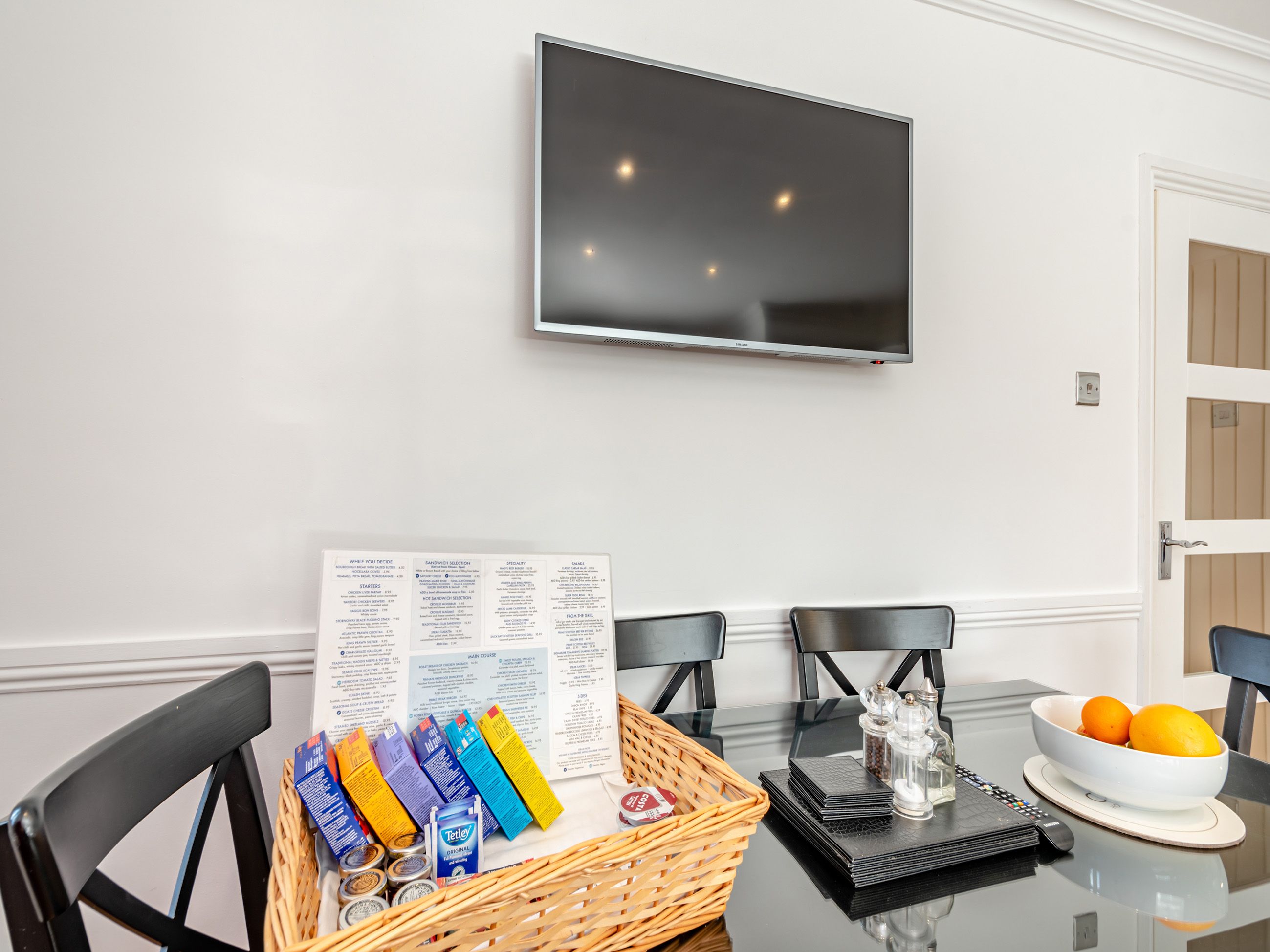 A dining area with a glass table, black chairs, a TV mounted on the wall, and a menu on the table.