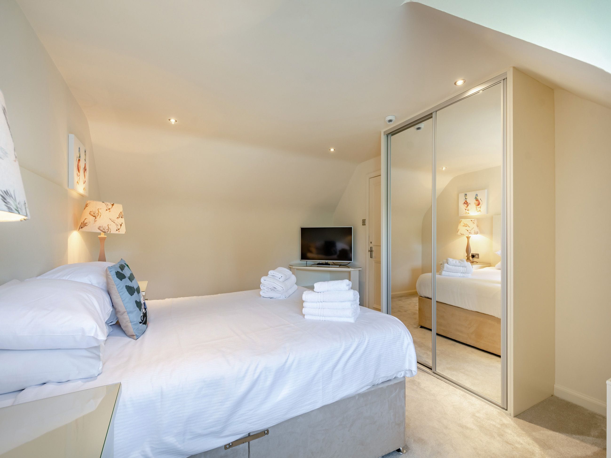 A modern hotel room with a bed, a TV, a glass door closet, and a wall-mounted lamp.