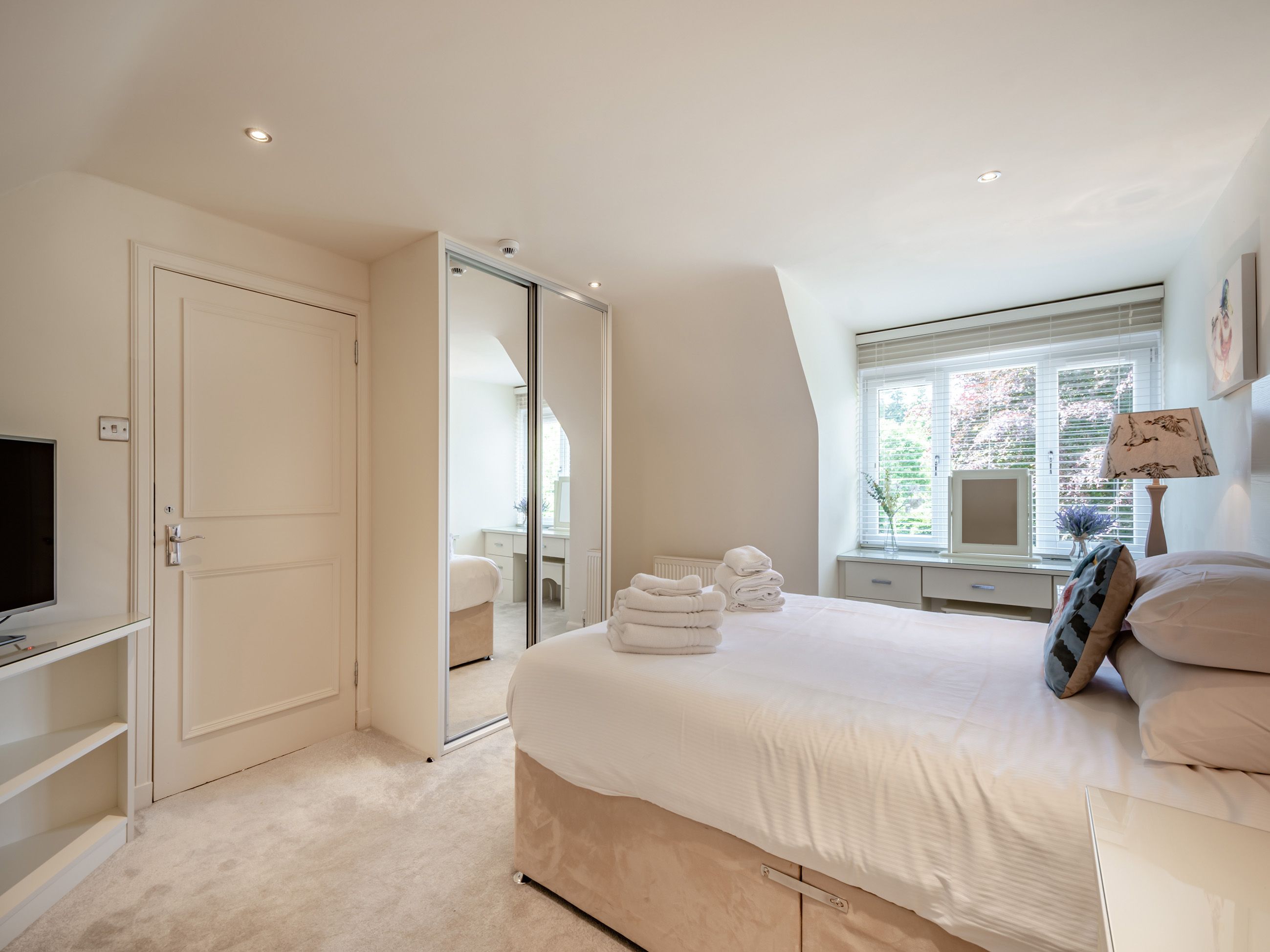 A bedroom with a bed, a TV, a desk, a window with blinds, and a mirror.