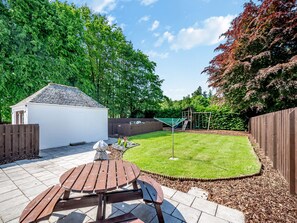 A backyard with a wooden table and benches, a small shed, a swing set, and a fenced area.