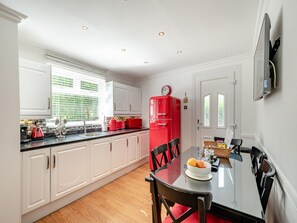 A modern kitchen with white cabinets, a red refrigerator, a dining table with chairs, and a window with blinds.