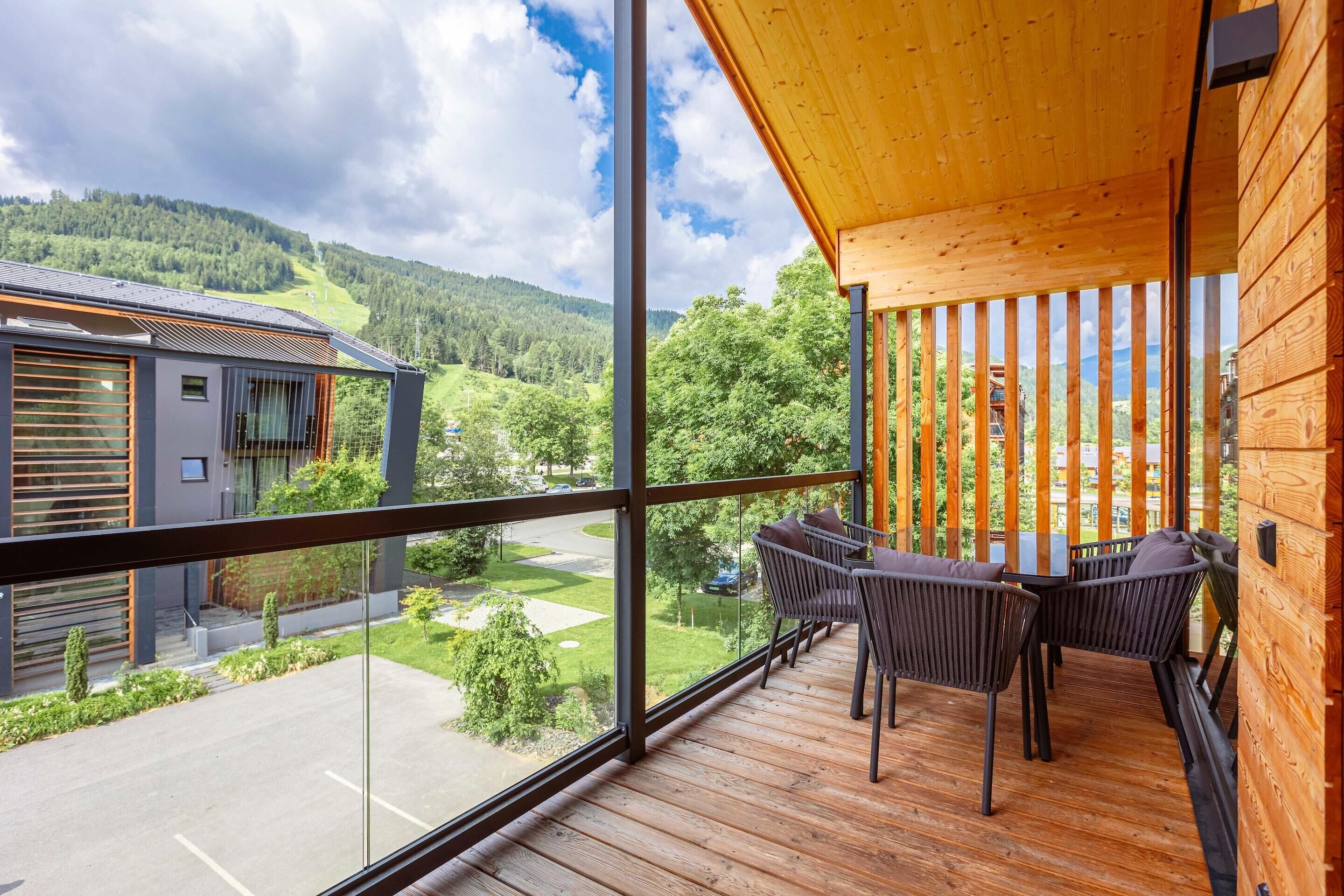 A balcony with a wooden floor and metal railing, overlooking a parking lot and a hilly landscape.