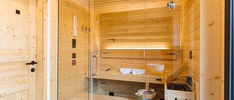 A sauna with wooden walls and a glass door, featuring a bench, towels, and a bucket.