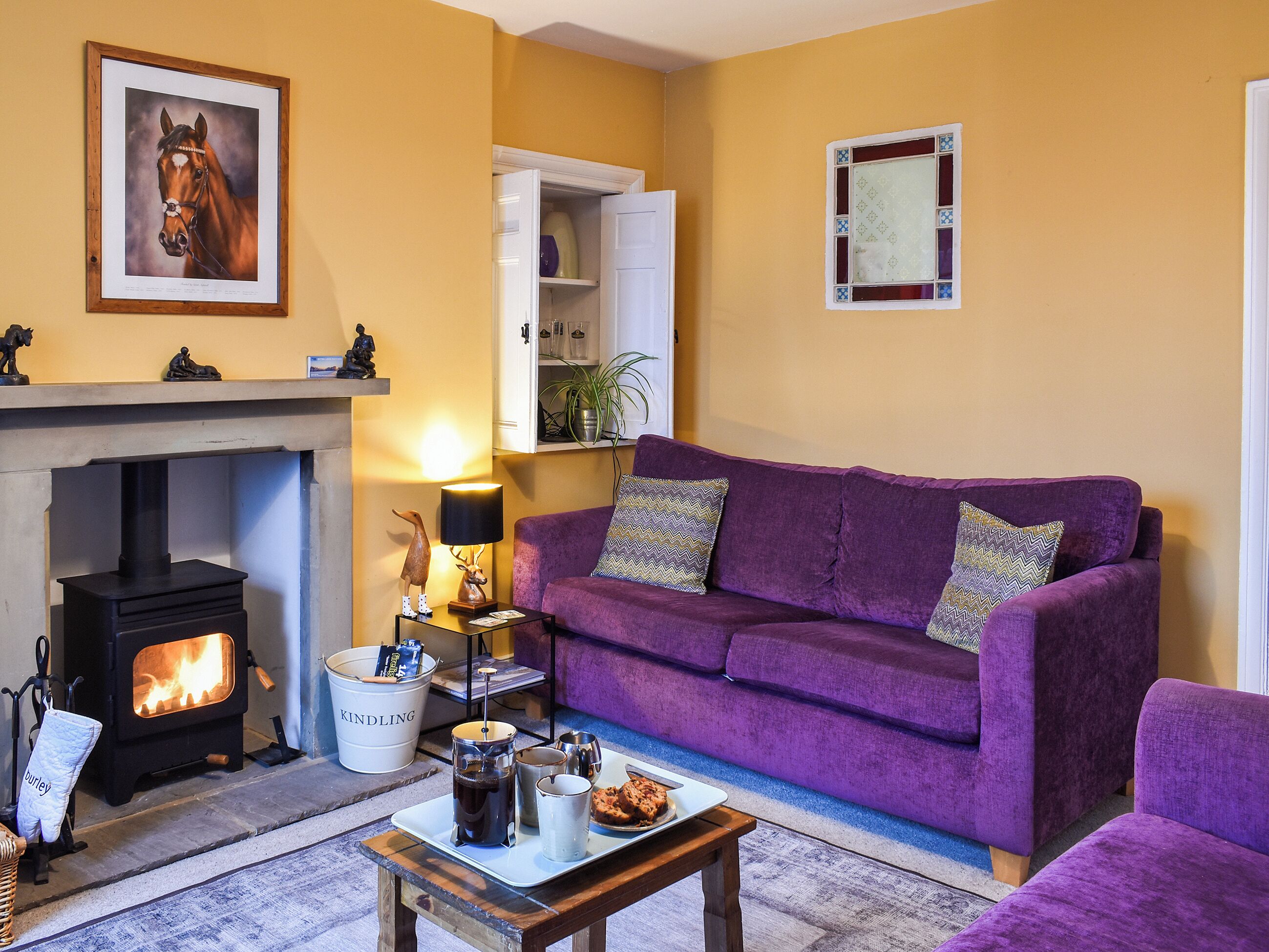 A cozy living room with a purple sofa, a fireplace, and a framed horse portrait.