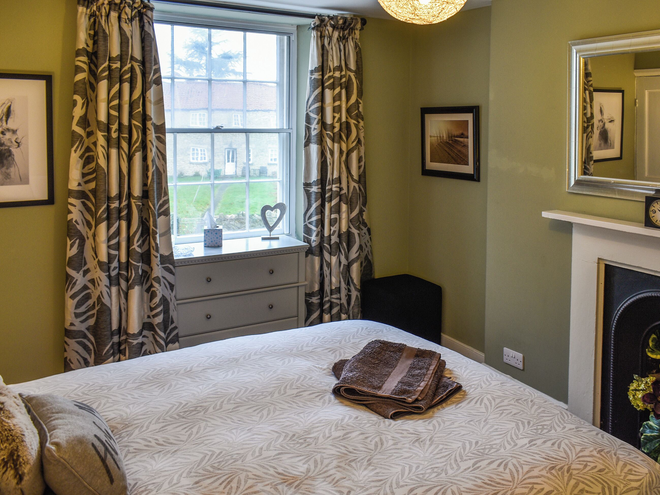 A bedroom with a bed, a dresser, a window with curtains, a fireplace, and framed pictures on the wall.