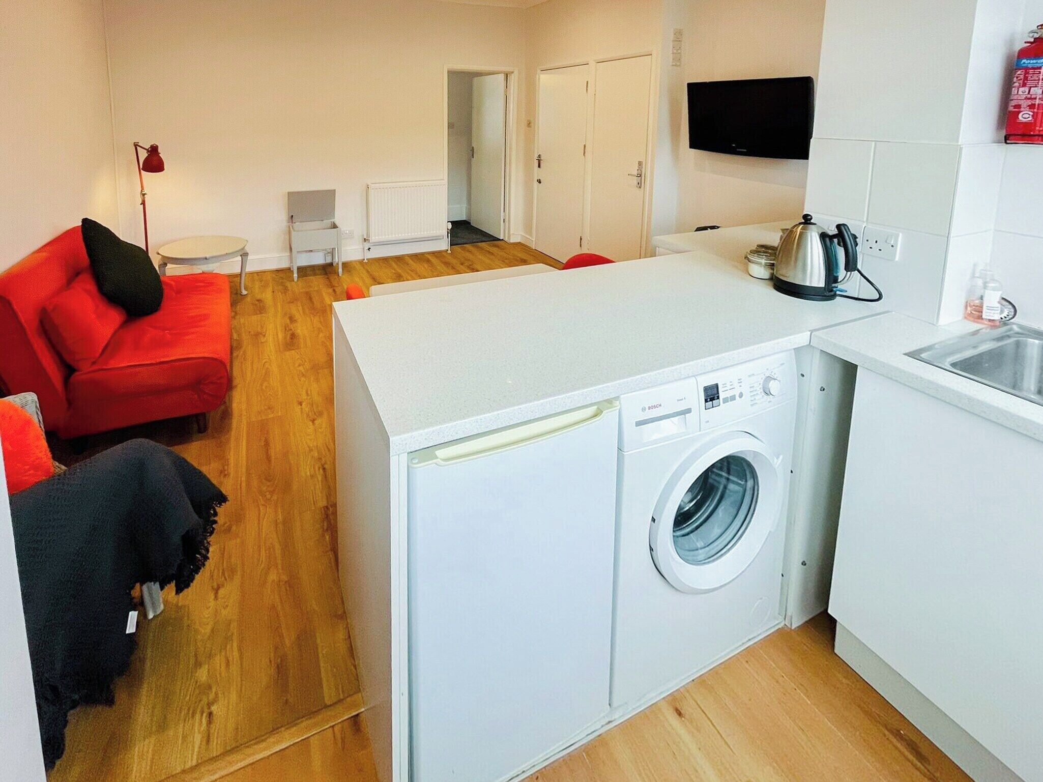 A compact apartment with a washing machine, a small kitchen area, a red sofa, and a television.