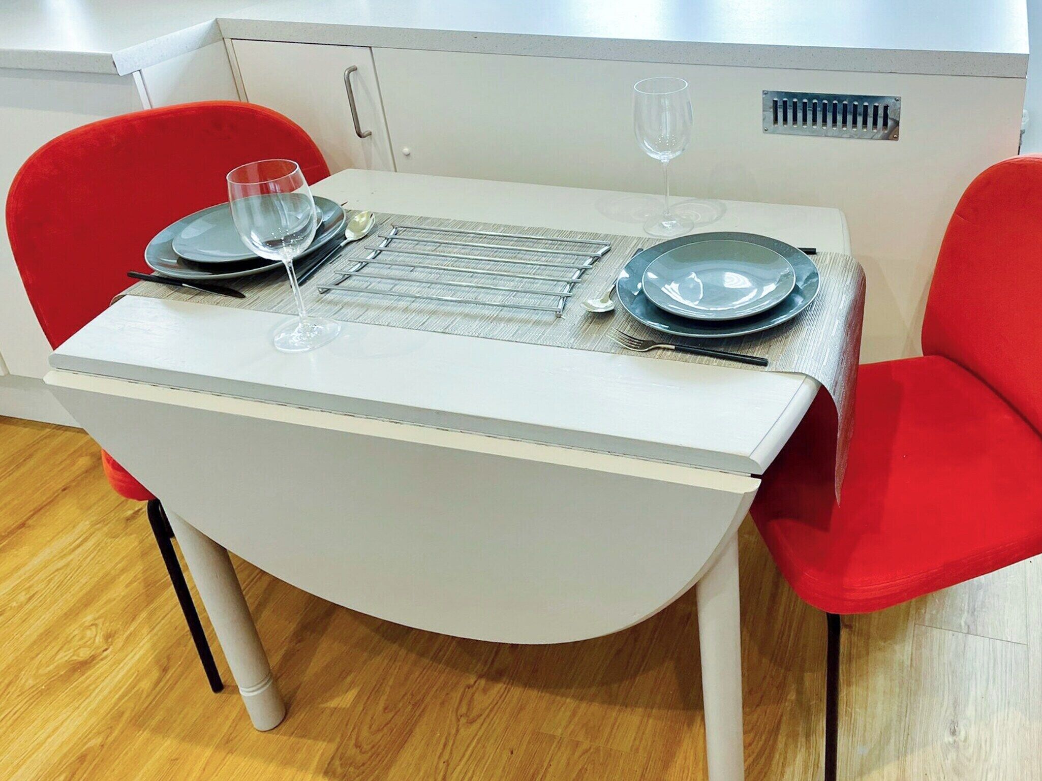 Uma mesa de jantar com estrutura metálica, duas cadeiras vermelhas e toalha branca.