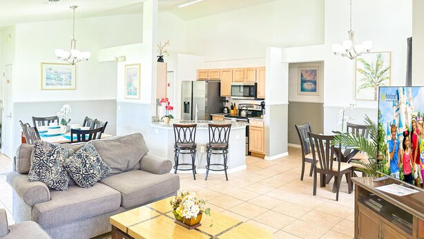 A spacious living room with a gray sofa, a coffee table, and a dining area with chairs. A kitchen with wooden cabinets and a microwave is visible in the background. - 4 Bedroom villa - private pool & game room near Disney (Kissimmee)