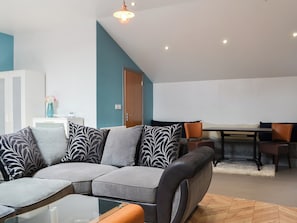 Un salon moderne comprenant un canapé d’angle, une table basse en verre, un coin repas avec une table et des chaises, et une porte donnant sur une autre pièce.