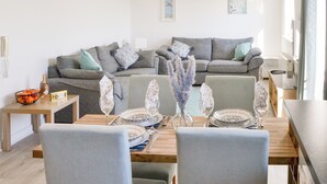 A modern living room with a dining area. There is a grey sofa set with patterned cushions, a wooden dining table set with plates, glasses, and cutlery. A vase with purple flowers is on the table.