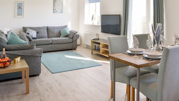 A modern living room with a grey sofa, a wooden dining table set for two, and a television mounted on the wall.