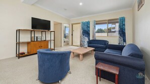 A living room with a sofa, a coffee table, a TV on a stand, and a bookshelf. - Aqua Sands Chalets-Nestled in the heart of Lancelin (Lancelin)