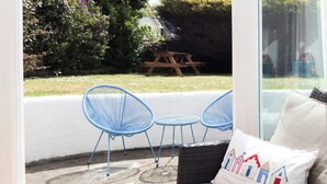 A room with a glass door leading to a patio with a table and chairs, a wicker sofa with a cushion, and a view of the garden. - 2 bedroom accommodation in Bude (Bude)