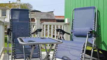 Two blue folding chairs with metal frames and a small table in between, positioned on a patio with a white fence and a green building in the background.