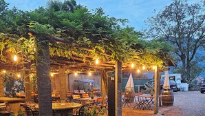 An outdoor restaurant with tables and chairs under a pergola, surrounded by greenery and gravel. - Francy (Ponte Capriasca)