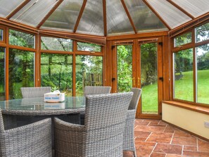 A conservatory with a glass table and wicker chairs, surrounded by large windows and a view of a garden.