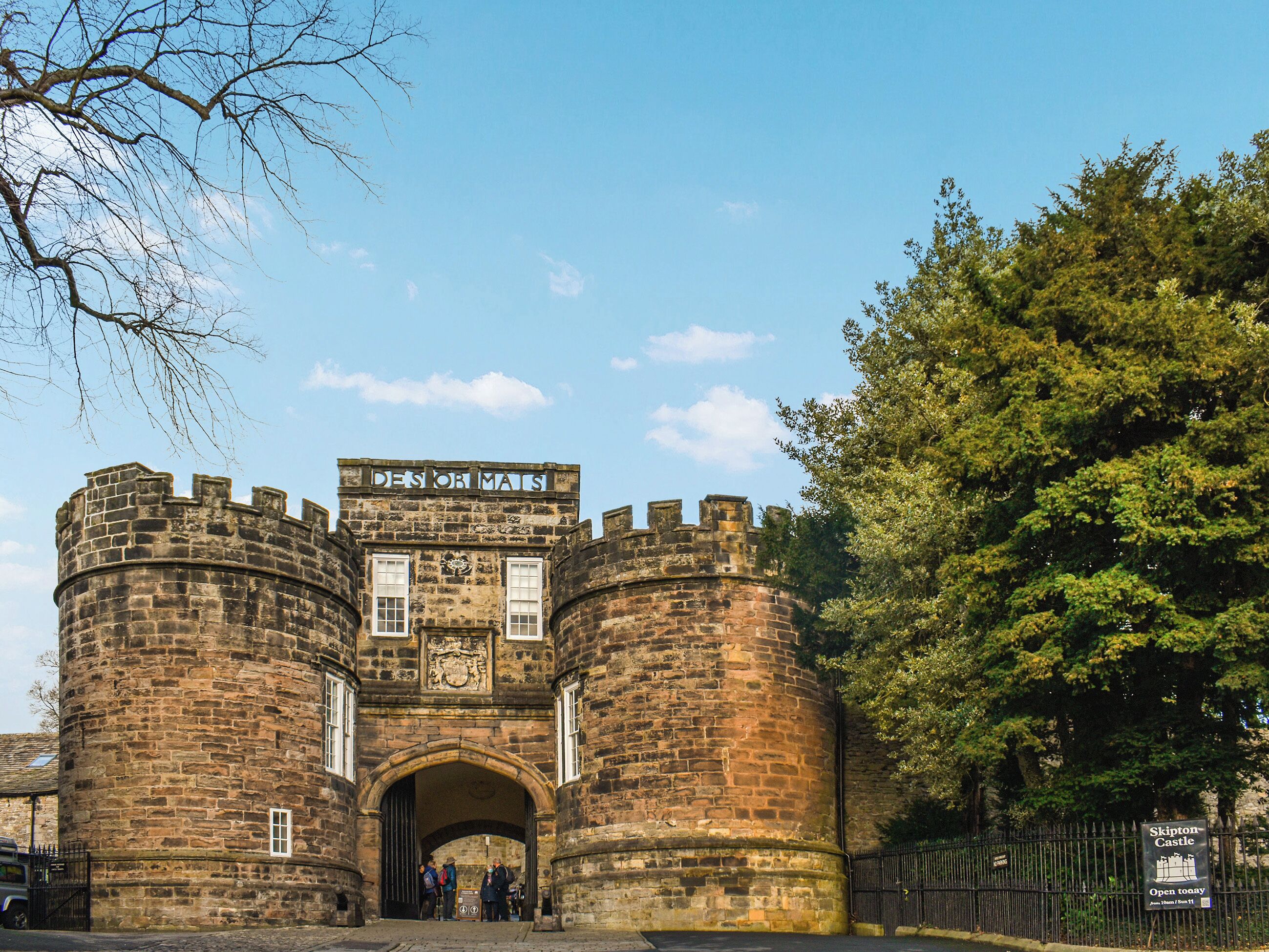 Una antigua puerta de piedra con un cartel que dice 'DEN OF MAUS' y un letrero de castillo que indica 'Skipton Castle Abierto Hoy'.
