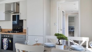 A modern kitchen with a stove, oven, and dishwasher. There is a dining area with a table set for two, a small potted plant, and a view into a bedroom with a bed and a nightstand. - 2 bedroom accommodation in Carnaby, near Bridlington (Carnaby, near Bridlington)