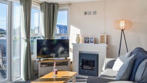 A modern living room with a sofa, a television, a fireplace, and a view of buildings outside. - 2 bedroom accommodation in Carnaby, near Bridlington (Carnaby, near Bridlington)