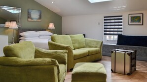 Une pièce comprenant un lit, deux fauteuils verts, un petit pouf, une valise, une lucarne, une fenêtre avec des stores, une lampe, un tableau et une carte accrochée au mur.