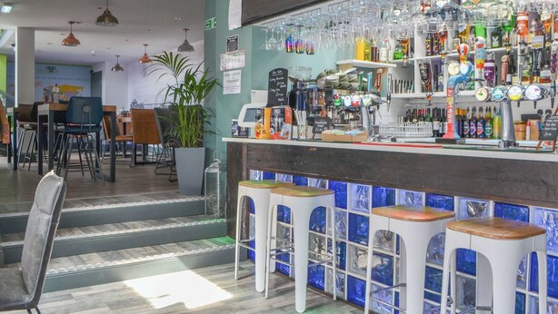 Un espace bar avec des tabourets, un comptoir où sont exposées différentes bouteilles et différents verres, et un coin salon avec des tables et des chaises.