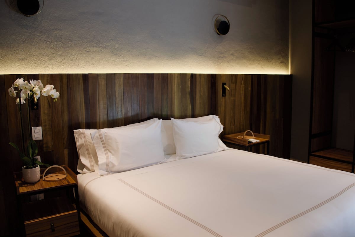 Een net opgemaakt bed met witte lakens, een houten hoofdbord en een klein nachtkastje met een lamp.