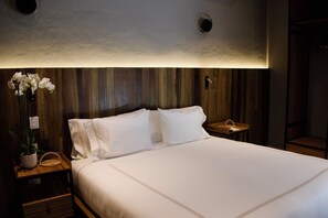 Un lit bien fait, avec du linge de lit blanc, une tête de lit en bois et une petite table de chevet sur laquelle se trouve une lampe.