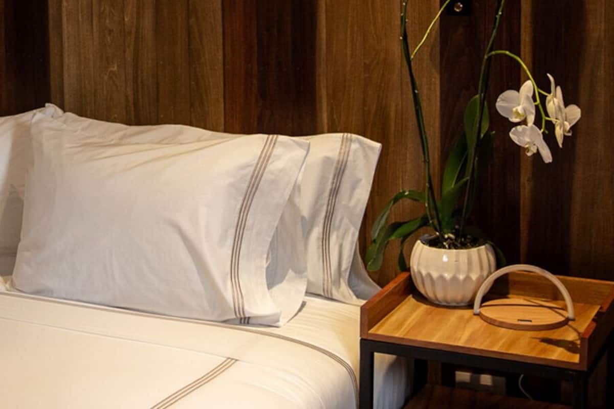 Een net opgemaakt bed met witte lakens, een houten nachtkastje met een lamp en een potorchidee op het nachtkastje.