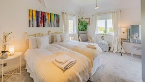 Une chambre spacieuse avec un grand lit, un bureau et une chaise. Il y a une fenêtre avec des rideaux, une plante et un tableau au mur.
