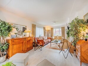 Uma sala de estar espaçosa com um armário de madeira, um sofá, uma mesa de centro e uma poltrona. Há plantas em vasos e um espelho grande na parede.
