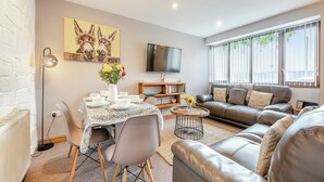 A modern living room with a dining area, a sofa set, a flat-screen TV, and a large window with blinds. - The Granary - UK41502 (Hundleton, near Pembroke)
