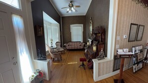 A spacious living room with hardwood floors, a ceiling fan, a sofa, armchairs, and a coffee table. There is a bookshelf with books and decorative items.