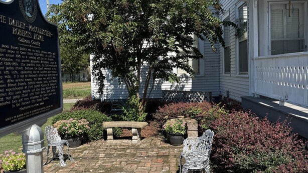 A historical marker sign, a brick pathway, a white house, and a garden with a bench and decorative chairs.