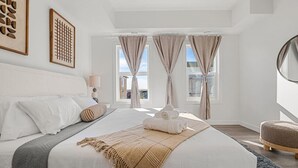 A modern bedroom with a large bed, two windows with curtains, a bedside lamp, and artwork on the wall. - Air Conditioned » Garage » 2BR Townhouse » Sleep 5 (Calgary)