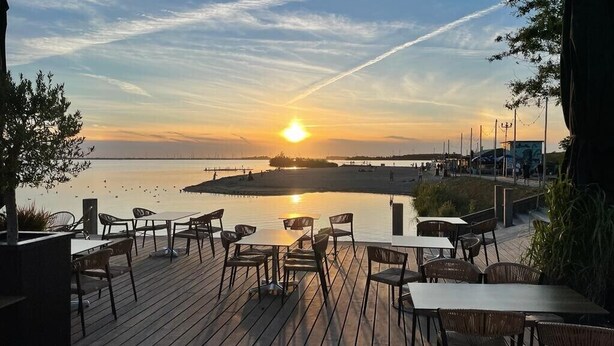 A wooden deck with tables and chairs, overlooking a calm body of water and a sunset. - Copenhagen Residence | Holiday home with Scandinavian flair | View of the water. (Harderwijk)