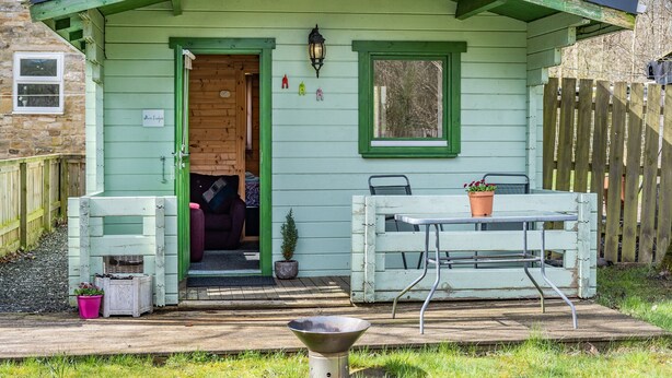Una pequeña estructura de madera verde con una puerta abierta a un cuarto, una ventana y una mesa con una planta en maceta.