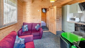 Un acogedor interior de cabaña con techo de madera, un conjunto de sofás, una zona de cocina y un televisor montado en la pared.